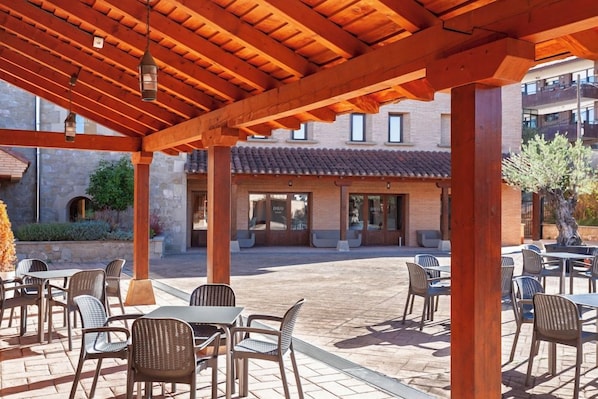 An outdoor patio area with tables and chairs, a covered wooden structure, and a building with a tiled roof in the background. - 4US Rioja Wine Hotel (Cenicero)