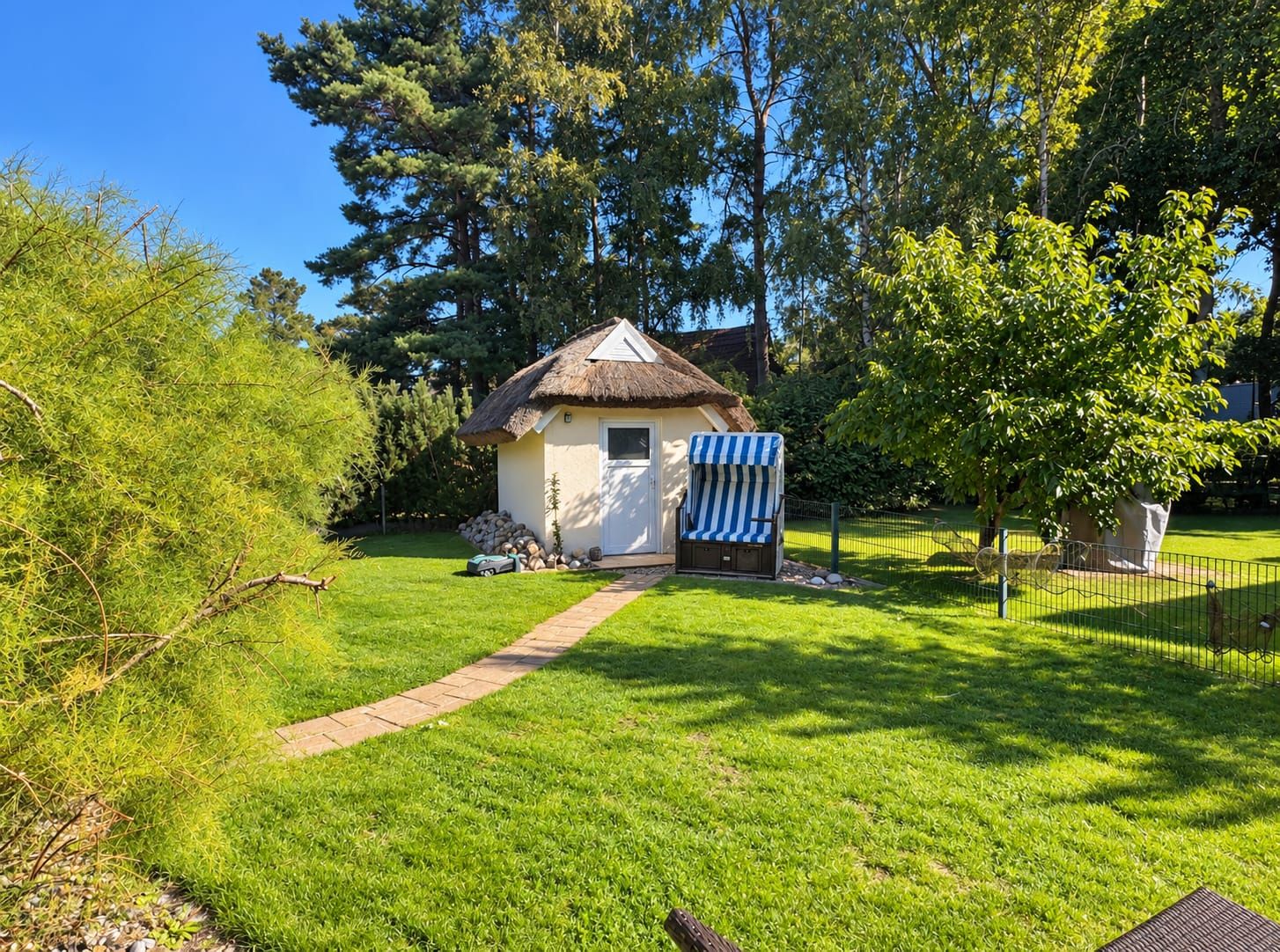 Ein kleines Haus mit einem Strohdach, einer überdachten Außen-Sitzgelegenheit und einem gepflasterten Weg zum Eingang.
