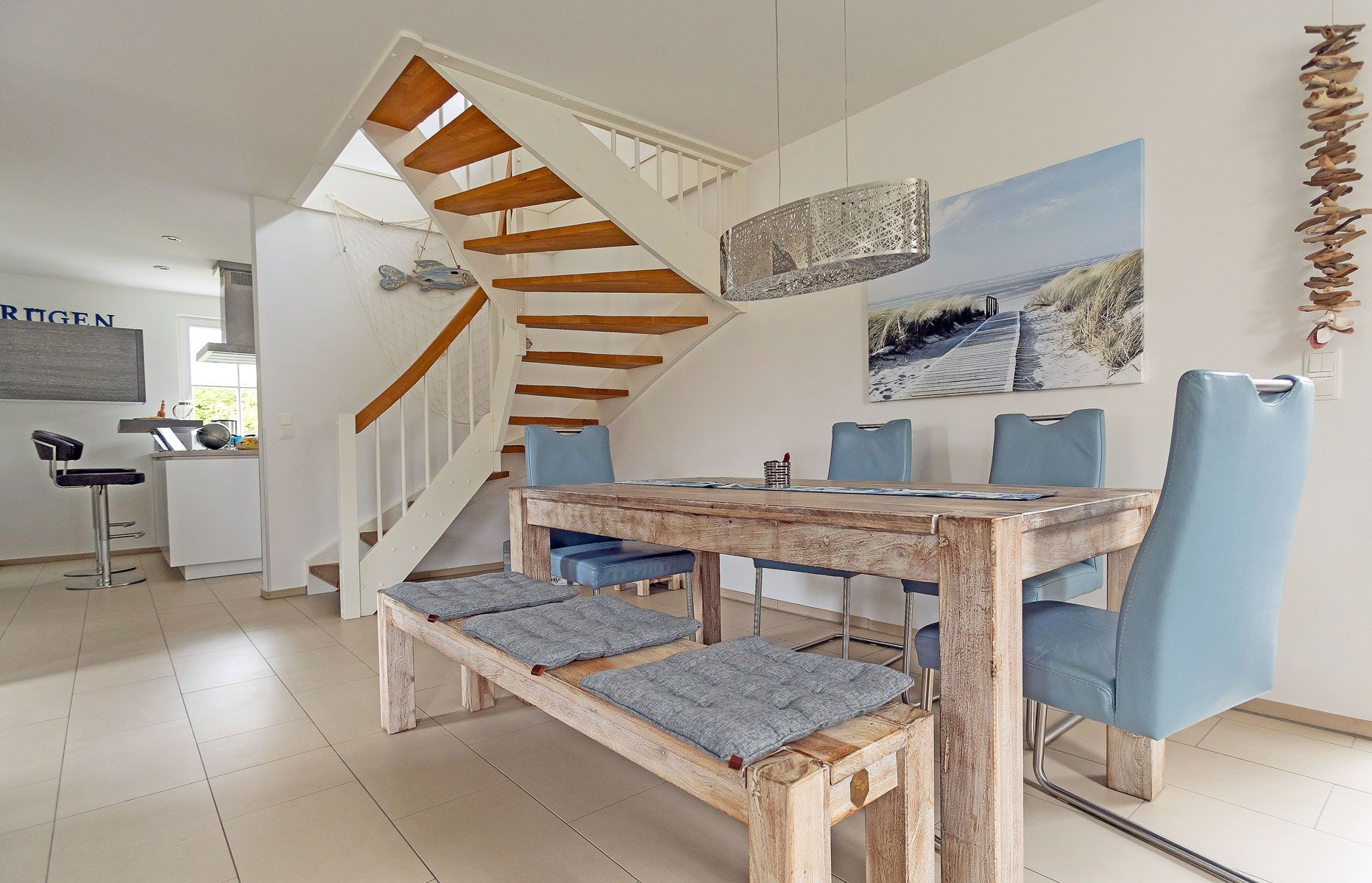Ein Essbereich mit Holztisch und blauen Stühlen, eine Treppe mit Holzstufen und im Hintergrund ein Küchenbereich.