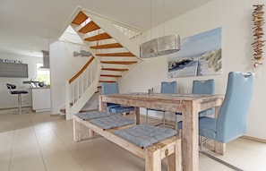 Ein Essbereich mit Holztisch und blauen Stühlen, eine Treppe mit Holzstufen und im Hintergrund ein Küchenbereich.