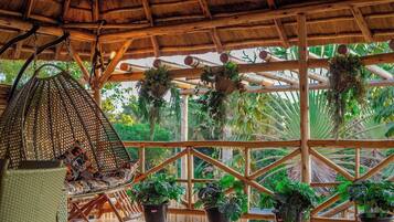 Una estructura de madera con techo de paja, plantas colgantes y una silla tejida.