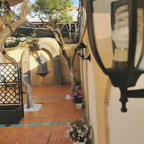 Un patio con suelo de baldosas, una reja metálica, un árbol y plantas en macetas.