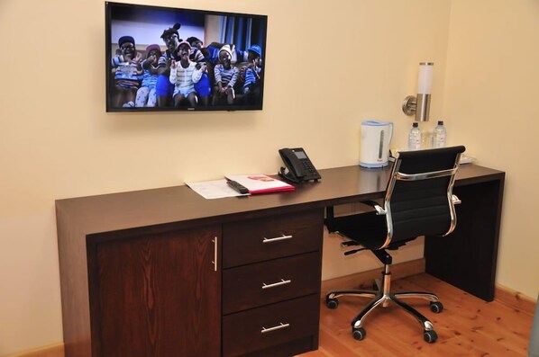 A hotel room with a wooden desk, a black office chair, a flat-screen TV mounted on the wall, a telephone, and a water dispenser. - Sigona Golf Club (Kikuyu)