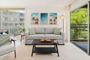 A modern living room with a grey sofa, a black coffee table, and a large window with a view of palm trees. - Hale Kamaole 244 - Modern Suite next to Beach (Kihei)