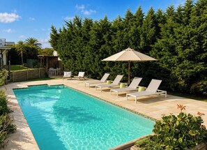 Ein rechteckiger Swimmingpool mit klarem, blauem Wasser, umgeben von einer gefliesten Liegefläche. Mehrere Liegestühle und ein einzelner Sonnenschirm sind vorhanden.
