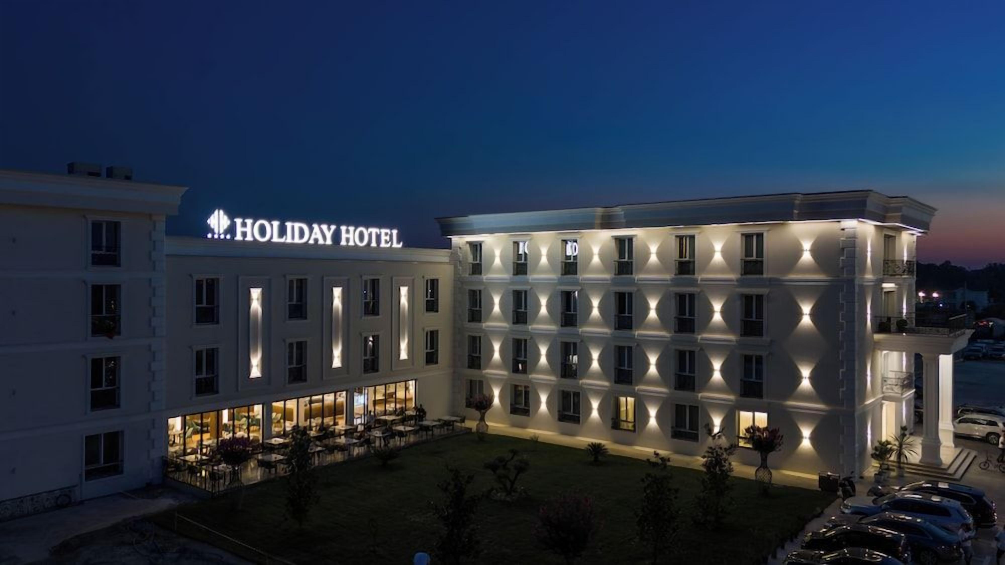 Un hotel de varios pisos con un llamativo letrero 'HOLIDAY HOTEL', exterior bien iluminado y un área de estacionamiento.