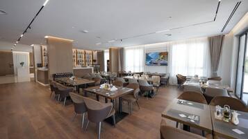 Un comedor de hotel moderno con varias mesas y sillas, una zona de bar y un ventanal con cortinas.