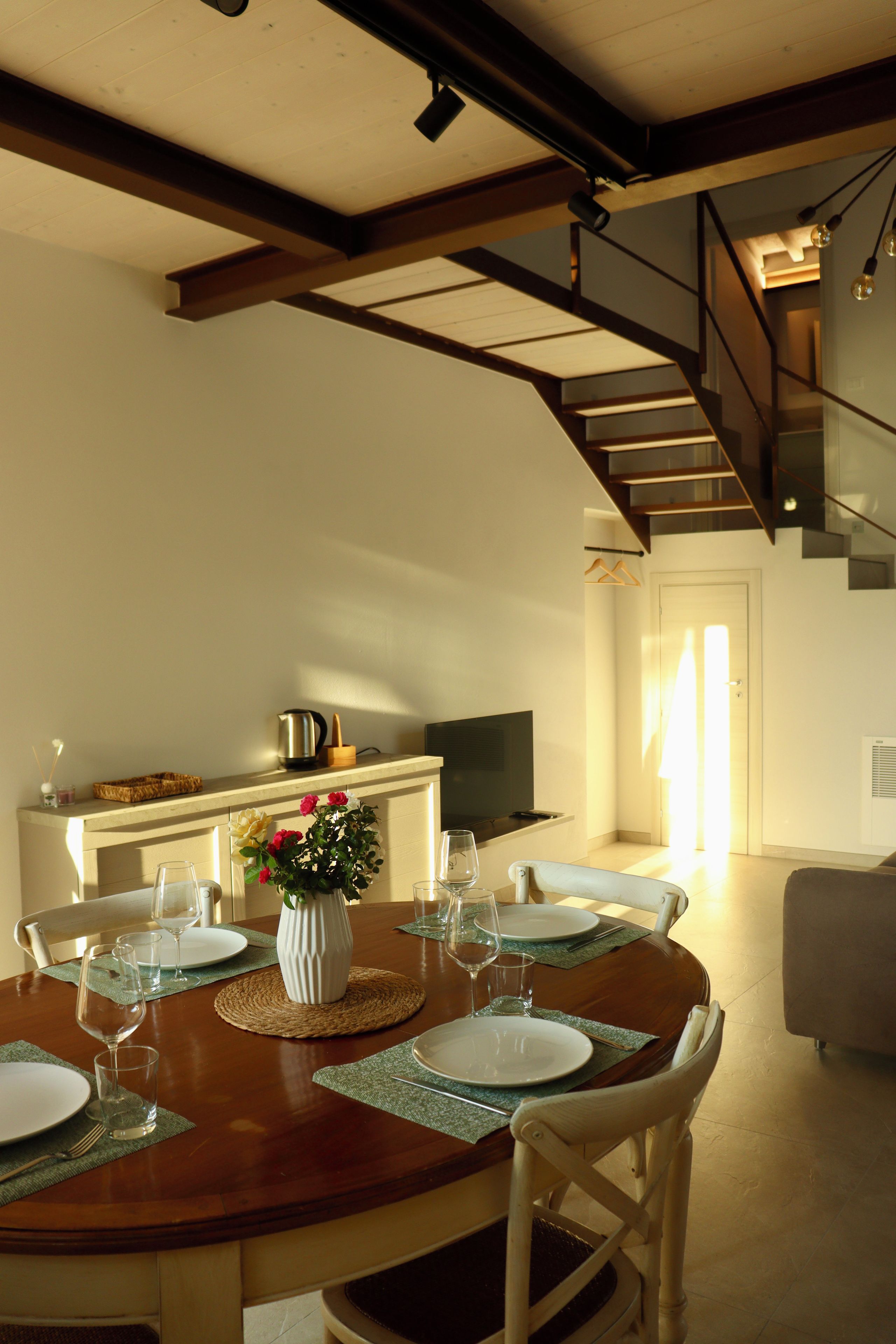 Un coin repas avec une table en bois dressée pour quatre personnes, un vase rempli de fleurs et un escalier en arrière-plan.