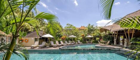 A swimming pool with lounge chairs and umbrellas, surrounded by tropical vegetation and a thatched-roof structure.