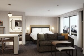 A hotel room with a bed, a sofa, a dining table, and a large window with a city view.