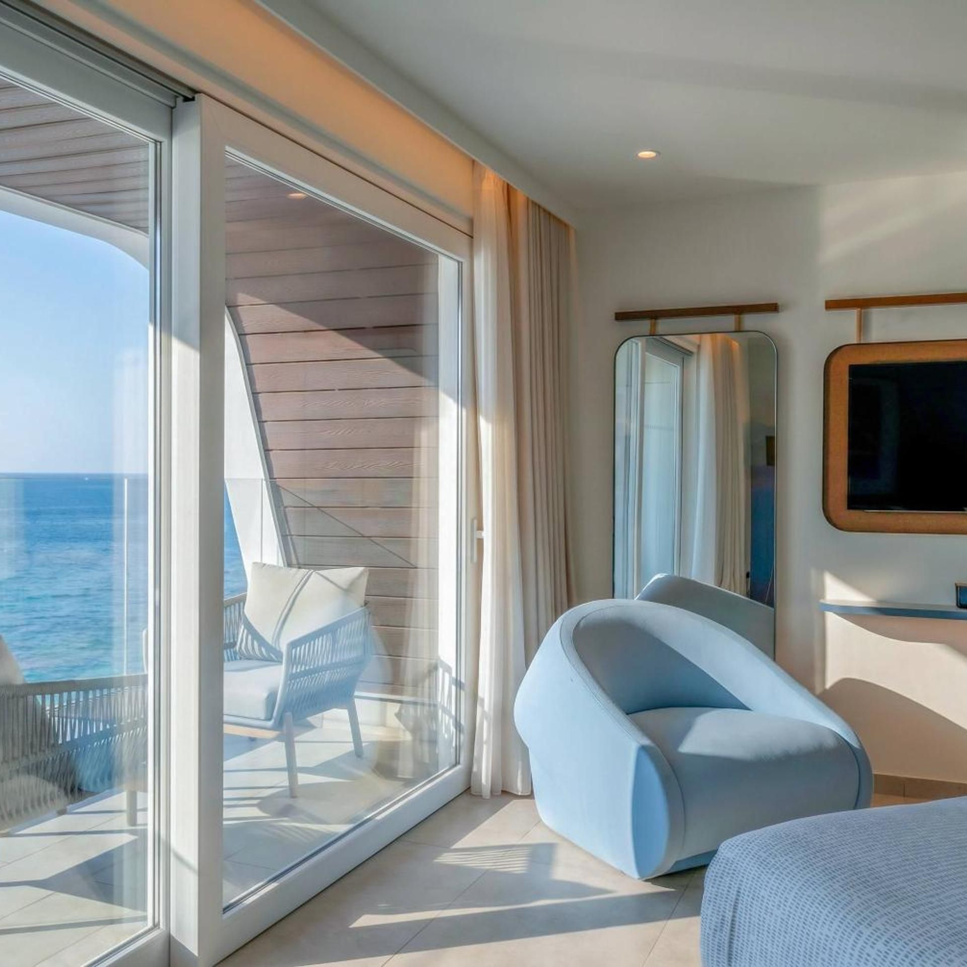 A modern room with a large sliding glass door leading to a balcony, a comfortable chair, a flat-screen TV mounted on the wall, and a bed.
