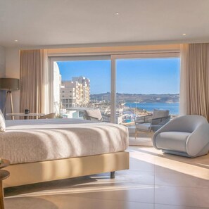 Une chambre d’hôtel avec un grand lit, un coin salon comprenant un fauteuil et une table, et une vue sur la mer et les bâtiments.