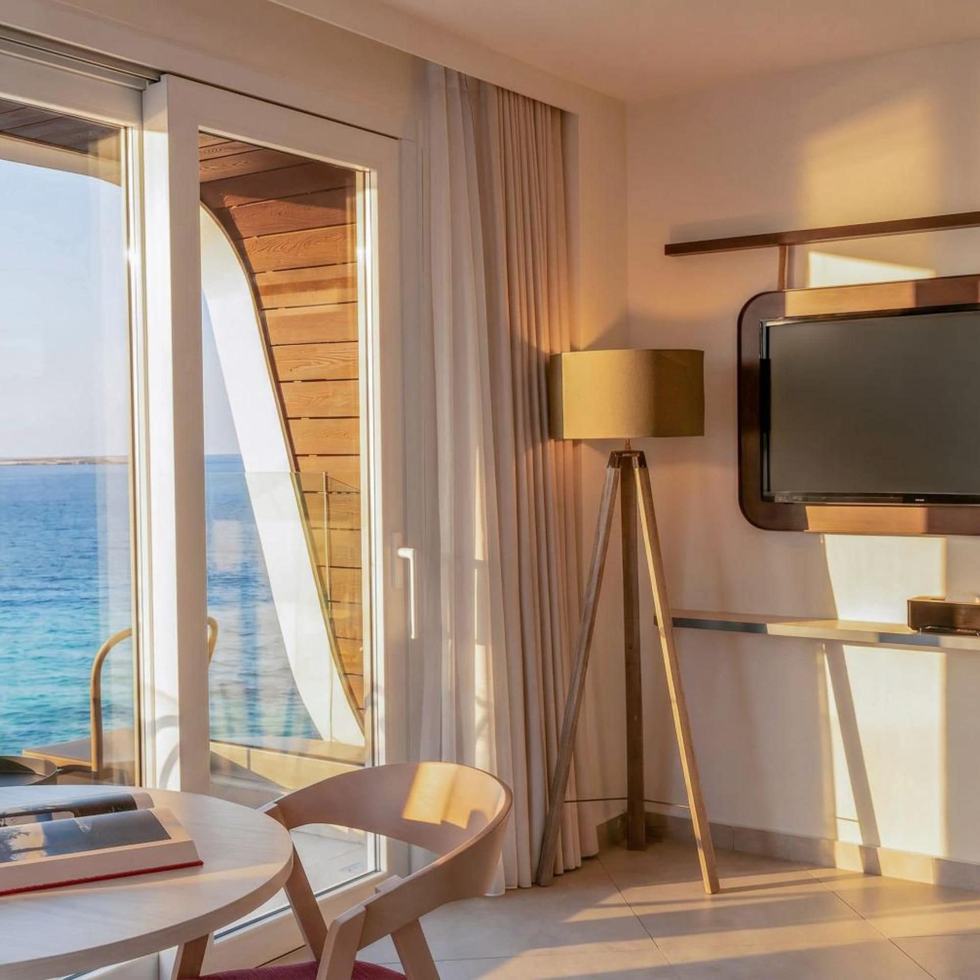 Quarto de hotel com uma janela ampla, televisão de tela plana, abajur e uma mesa com cadeira.