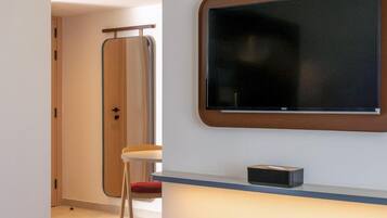 Quarto de hotel moderno com uma televisão de tela grande fixada na parede, uma pequena mesa com uma cadeira e uma porta que dá para outro quarto.