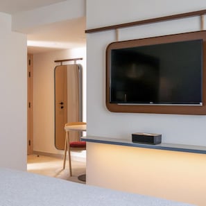 A modern hotel room with a large flat-screen TV mounted on the wall, a small table with a chair, and a door leading to another room.
