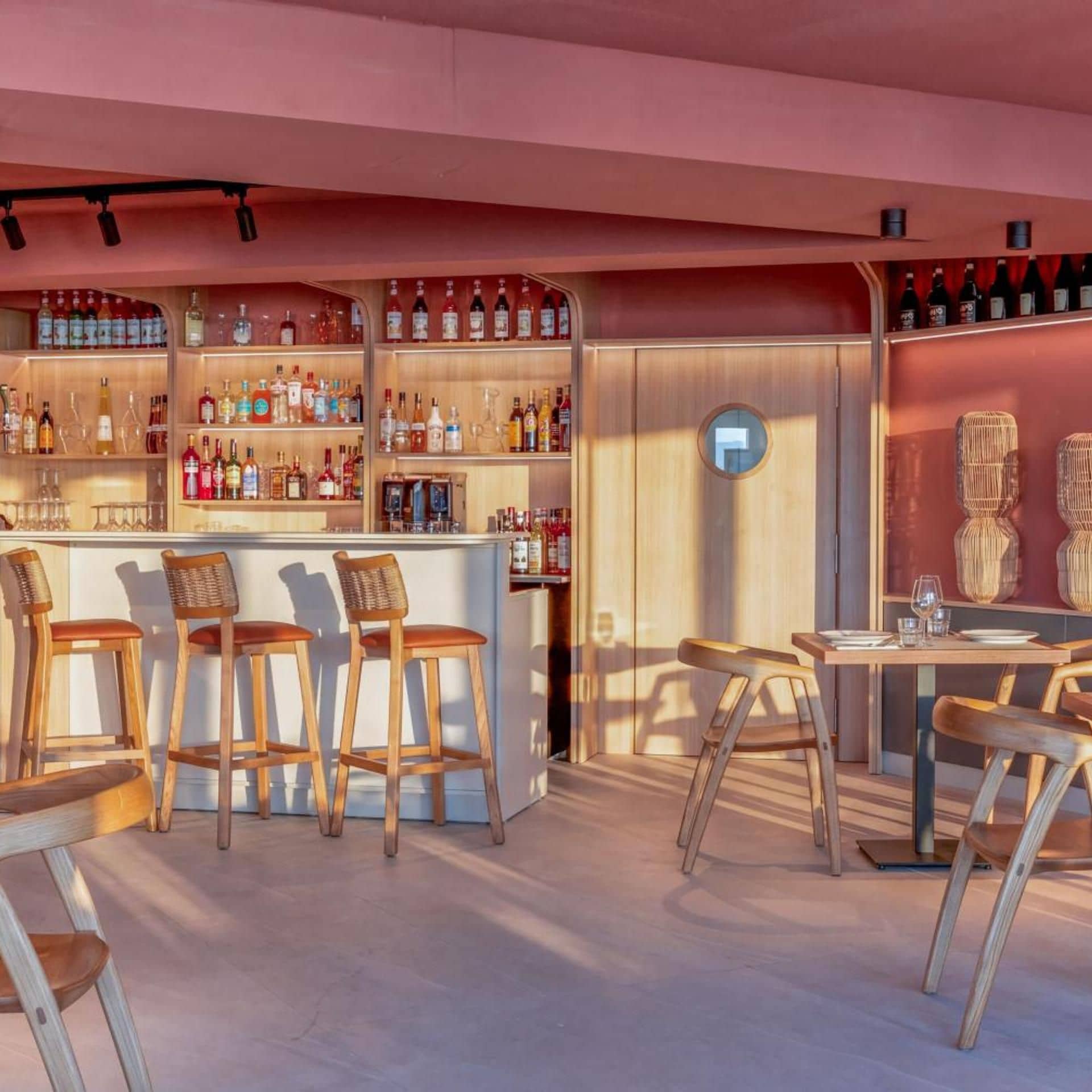 Uma área de bar com banquetas de madeira e balcão, prateleiras com garrafas e um espelho redondo na parede.