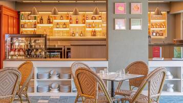 A bar area with shelves of bottles and glasses, wicker chairs around a table, and a tiled floor.