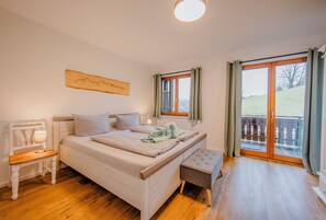 Quarto com cama, mesa de cabeceira, banco, janela e vista para uma varanda.