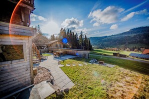 Eine Holzhütte mit Terrasse, einem kleinen Geräteschuppen und Blick auf eine hügelige Landschaft.