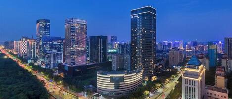Une vue d’ensemble d’une ville au crépuscule, avec des immeubles modernes de grande hauteur, un bâtiment aux lignes courbes doté d’une cour intérieure, et une route où circulent des véhicules.