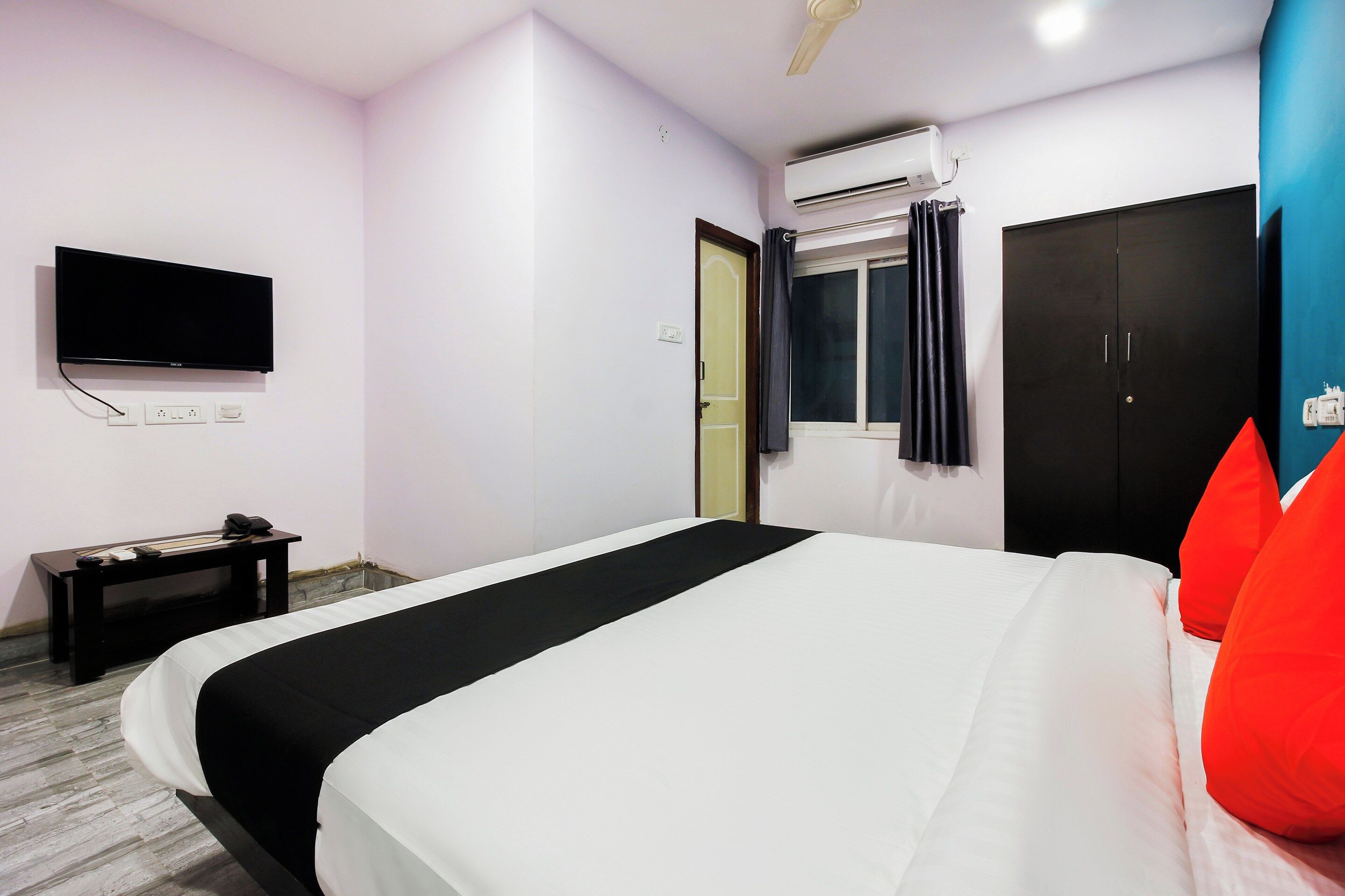 A hotel room with a bed, a flat-screen TV, an air conditioning unit, a desk with a phone, and a wardrobe.