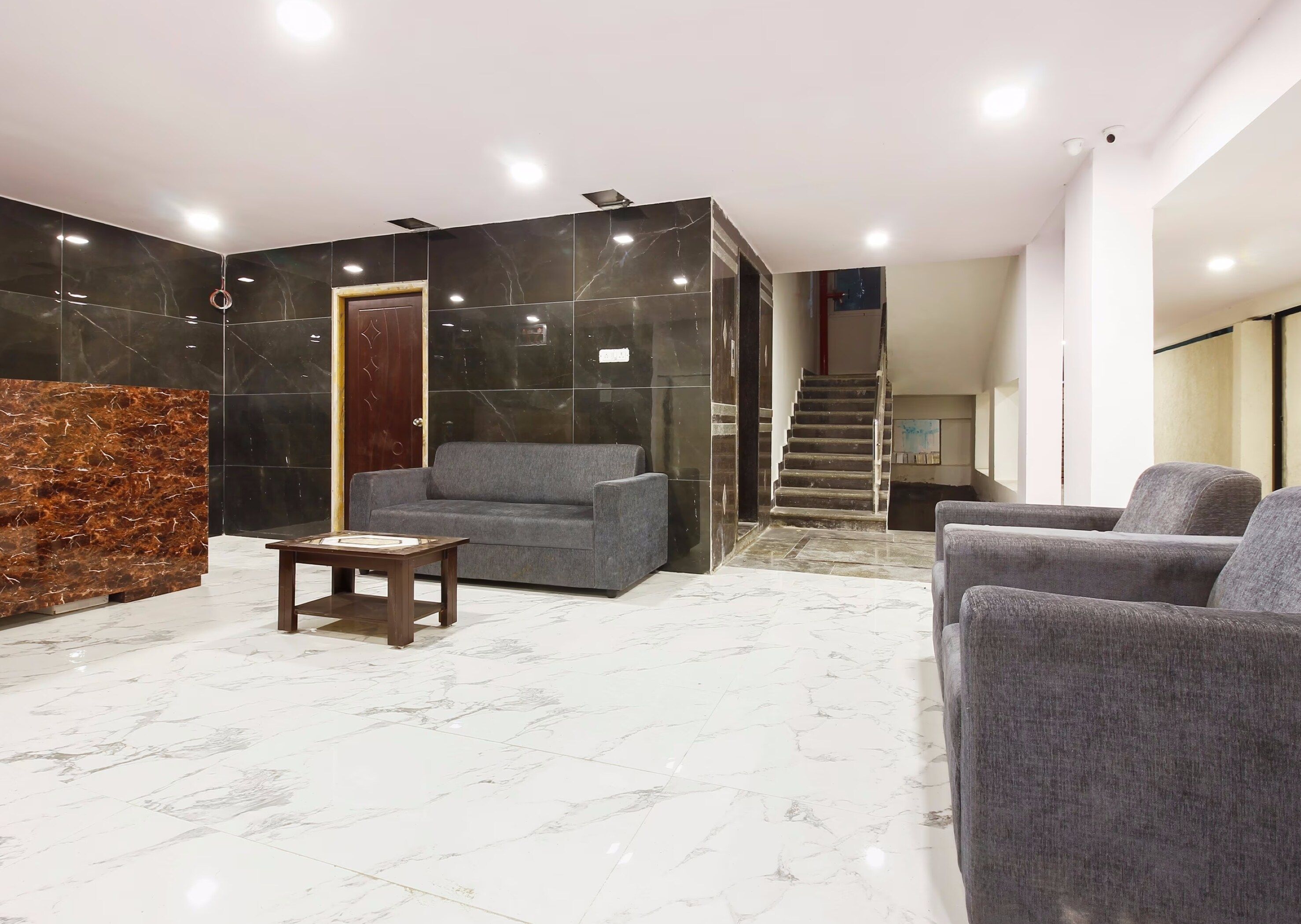 Vestíbulo con mostrador de recepción, sofás grises, una mesa de centro de madera y una escalera que conduce a un piso superior.