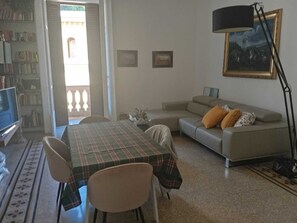 Ein Wohnzimmer mit Sofa, Tisch und Stühlen. Links befindet sich ein Fernseher und rechts eine Lampe.
