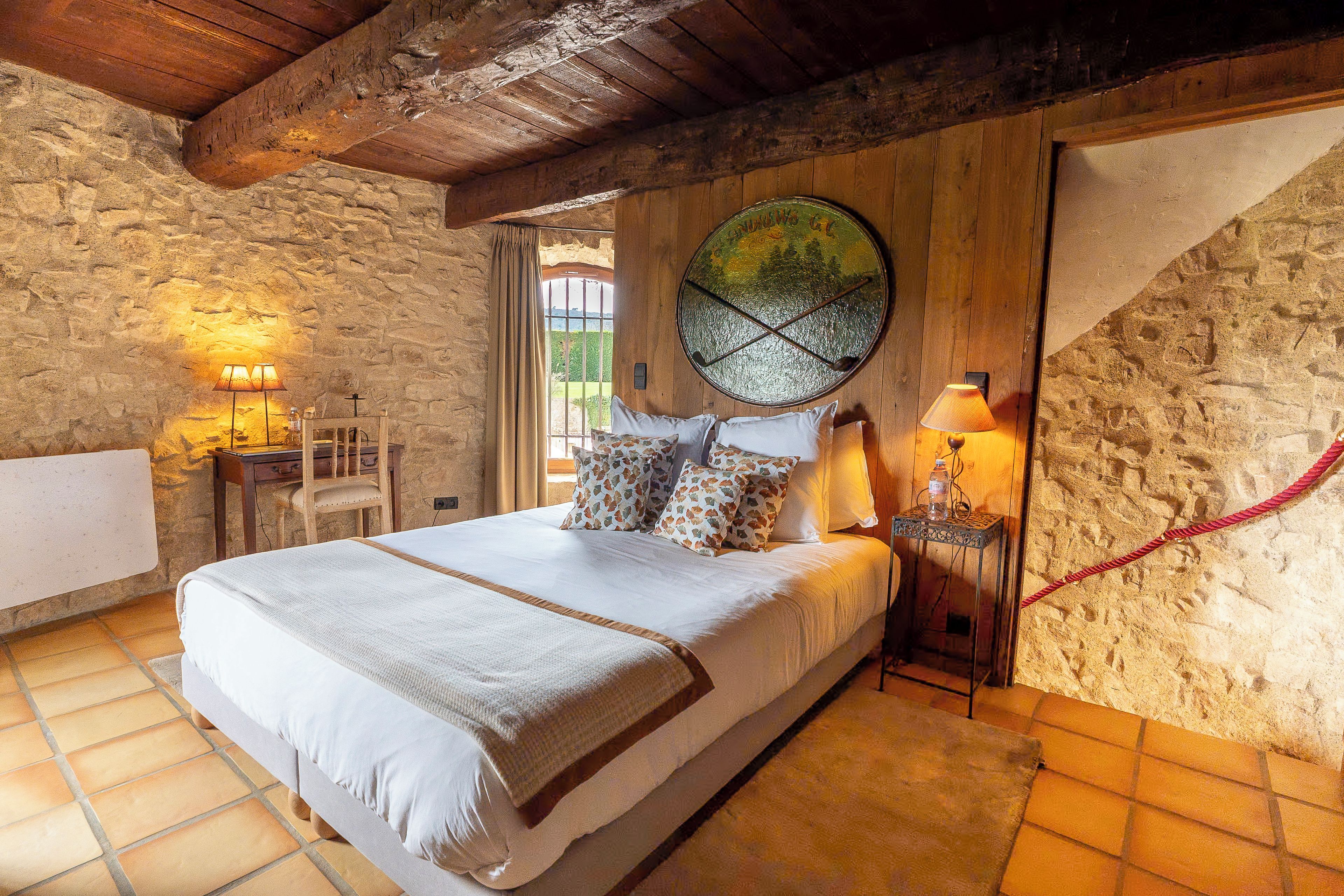 A bedroom with a stone wall, a bed with a white bedspread, a wooden headboard, and a large window with a view.