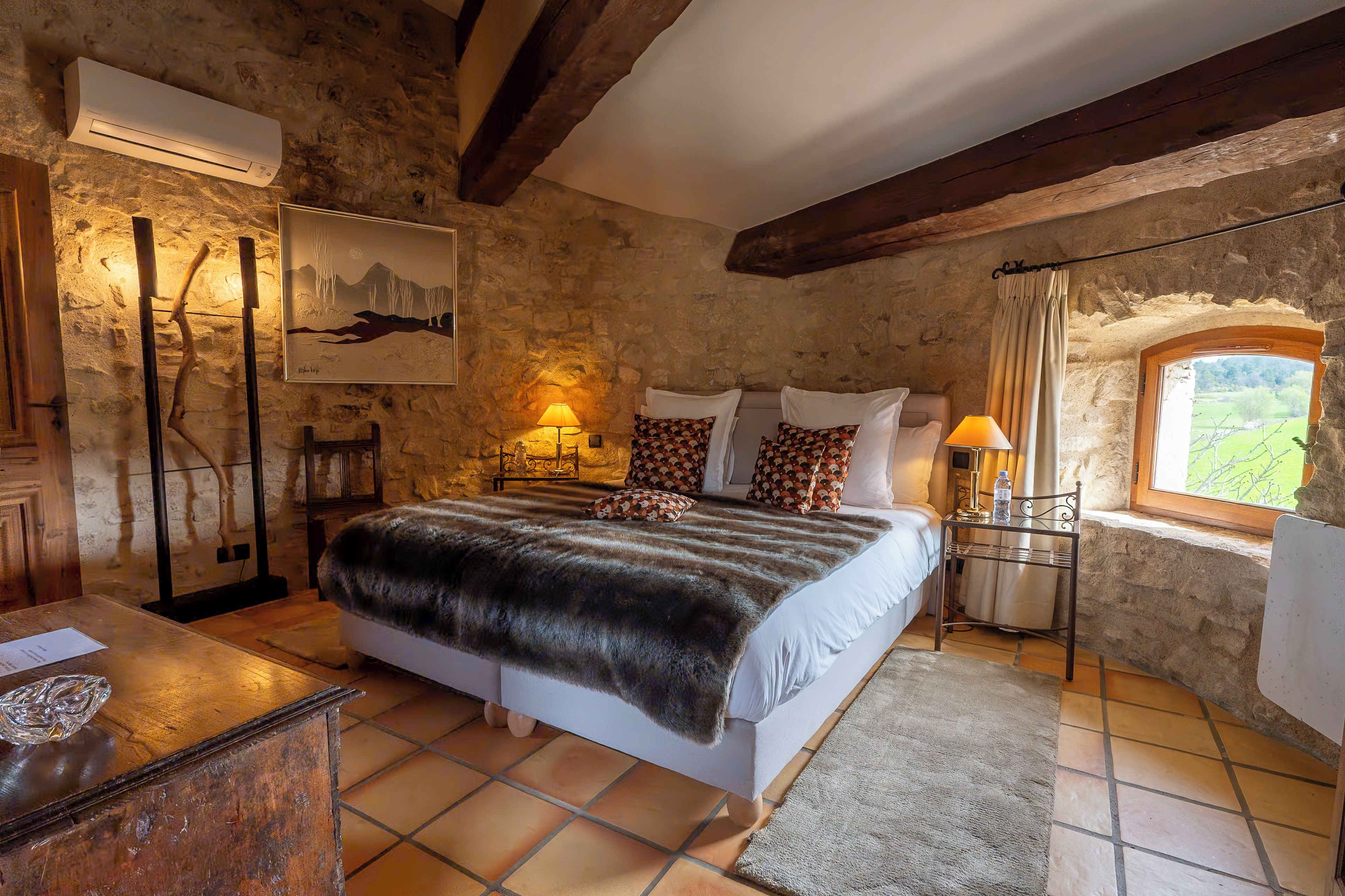 A rustic bedroom with a stone wall, a bed with patterned pillows, a wooden nightstand, a lamp, and a window with a view.