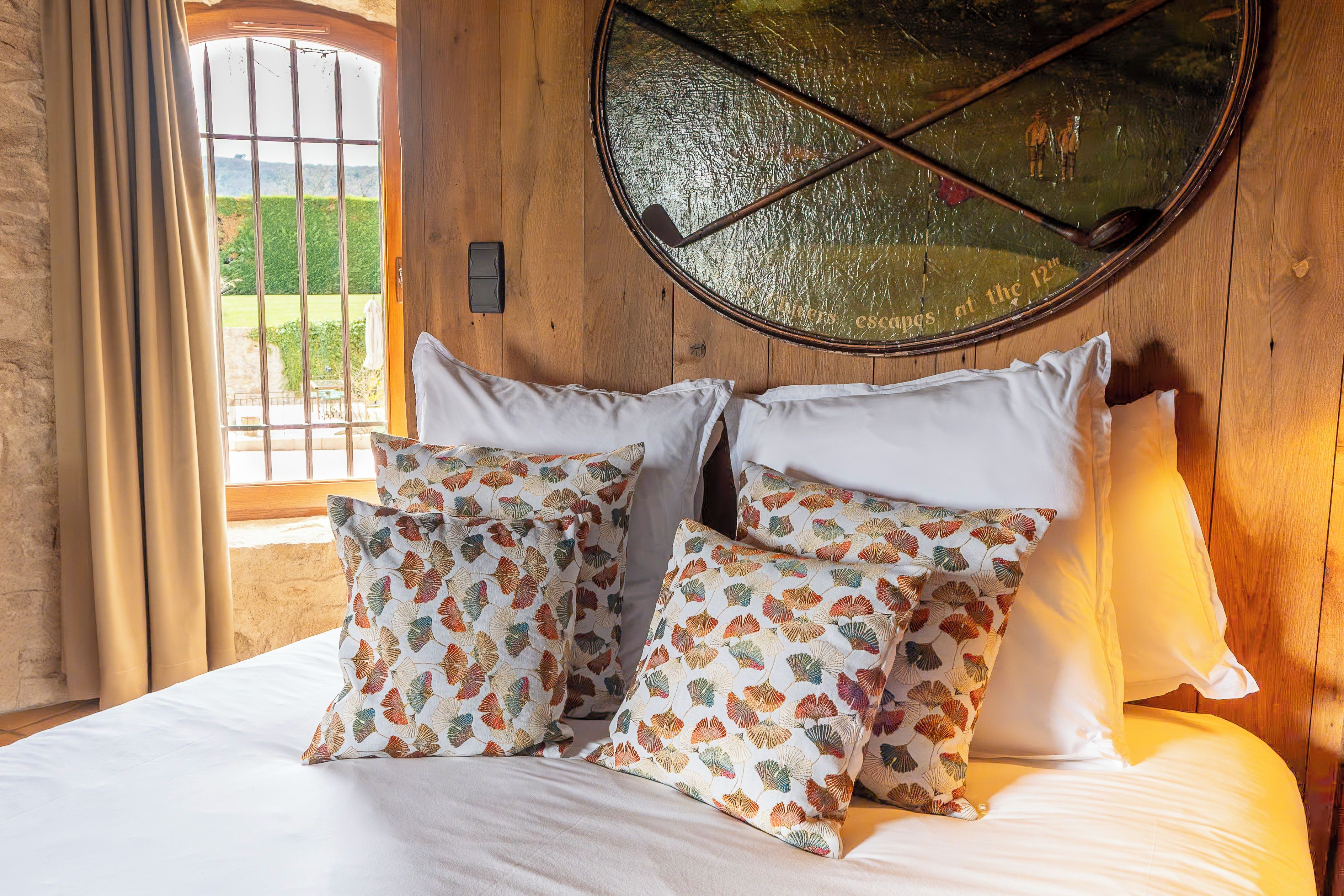 Un lit avec une literie blanche et des coussins à motifs, un miroir rond accroché au mur et une fenêtre avec des rideaux.