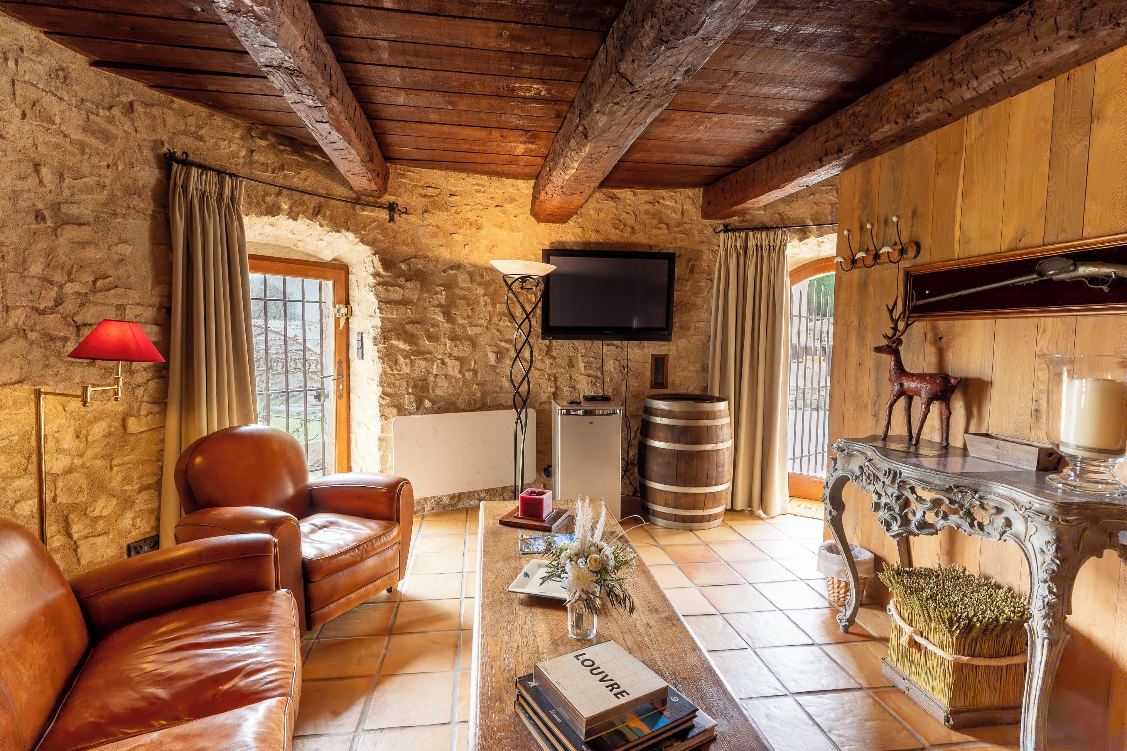 Una habitación rústica con paredes de piedra, techo de madera, sofá de cuero, televisor, chimenea con cabeza de venado y un barril.