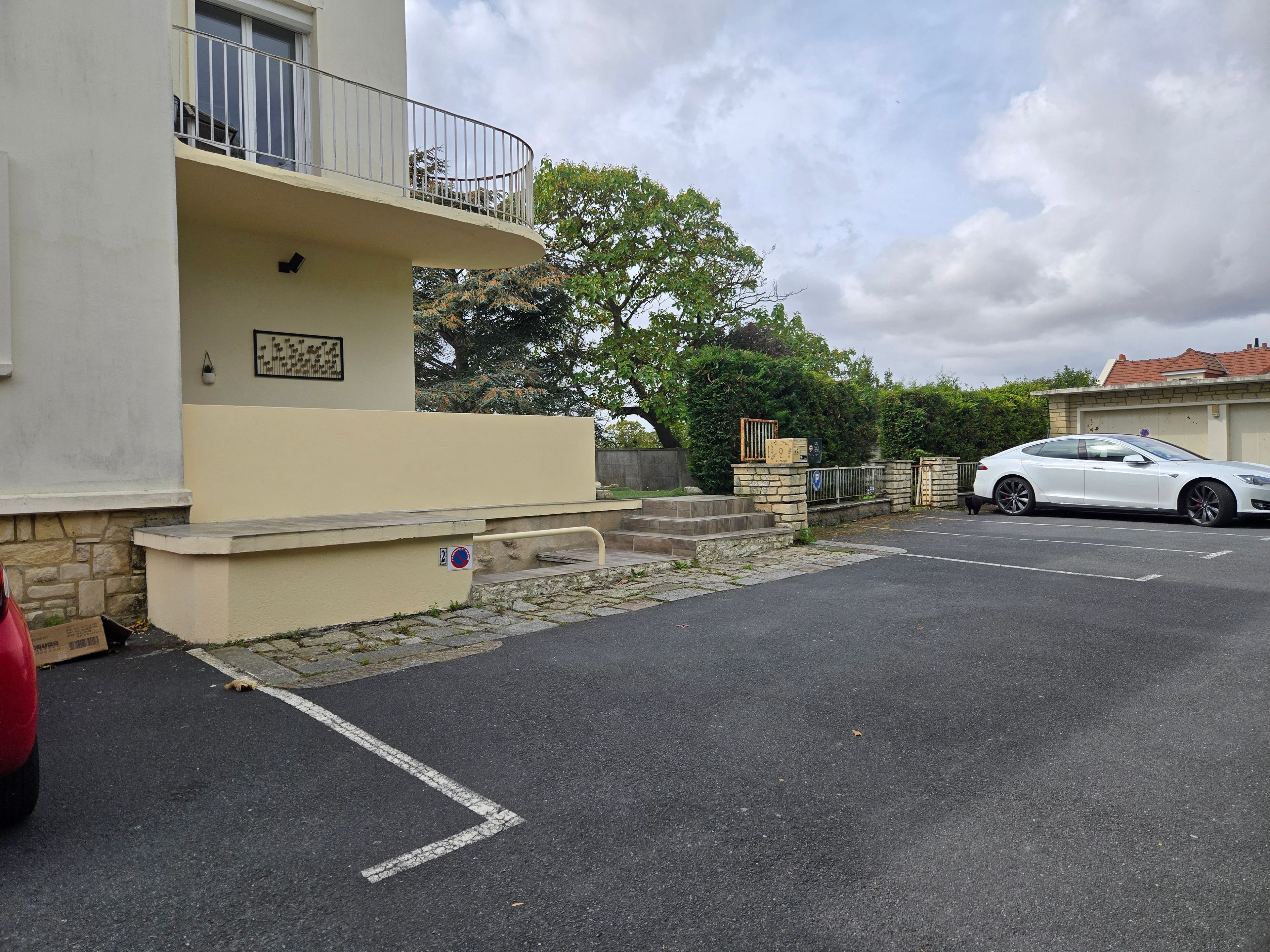 A residential building with a balcony, a parked white car, and a red car in the driveway.