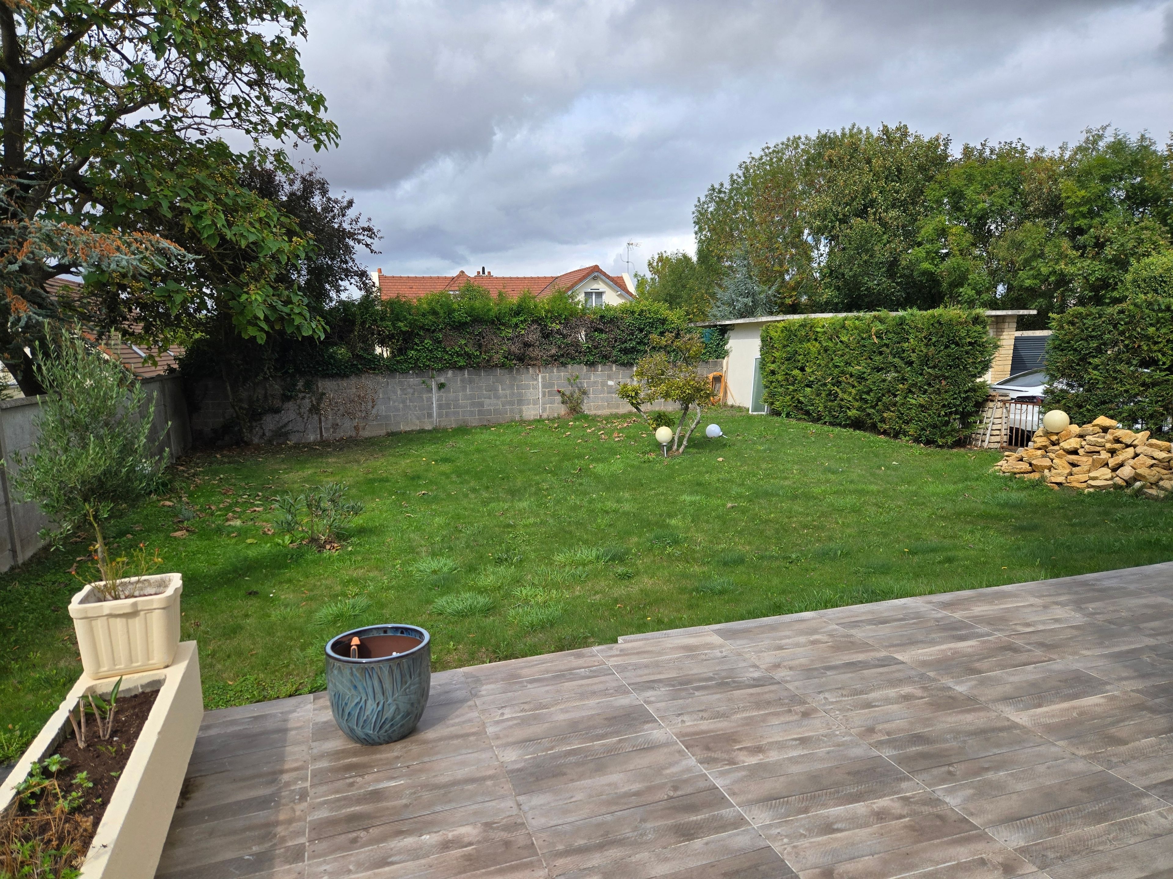 Un patio trasero con terraza de madera, un pequeño jardín con plantas y un muro de piedra.