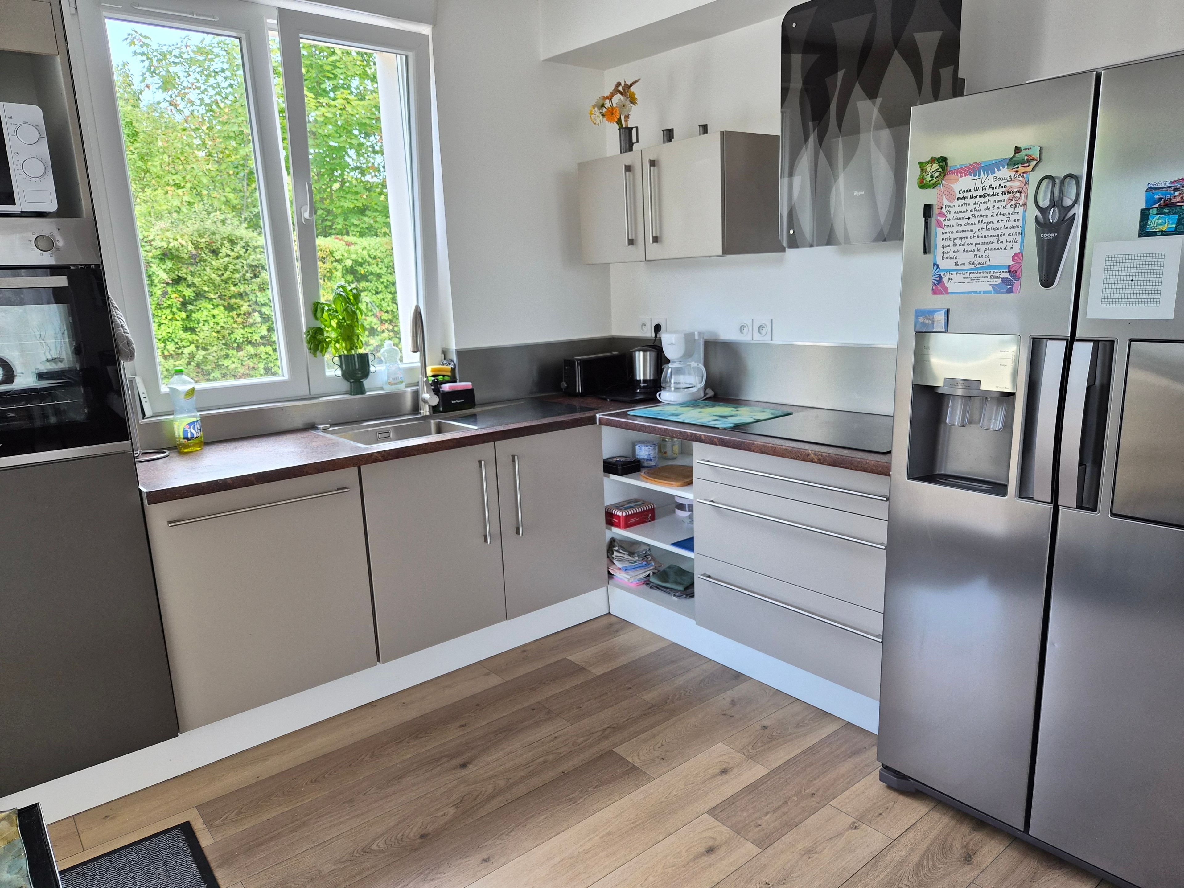 A modern kitchen with a stainless steel refrigerator, a built-in oven, and a window providing a view of greenery outside.