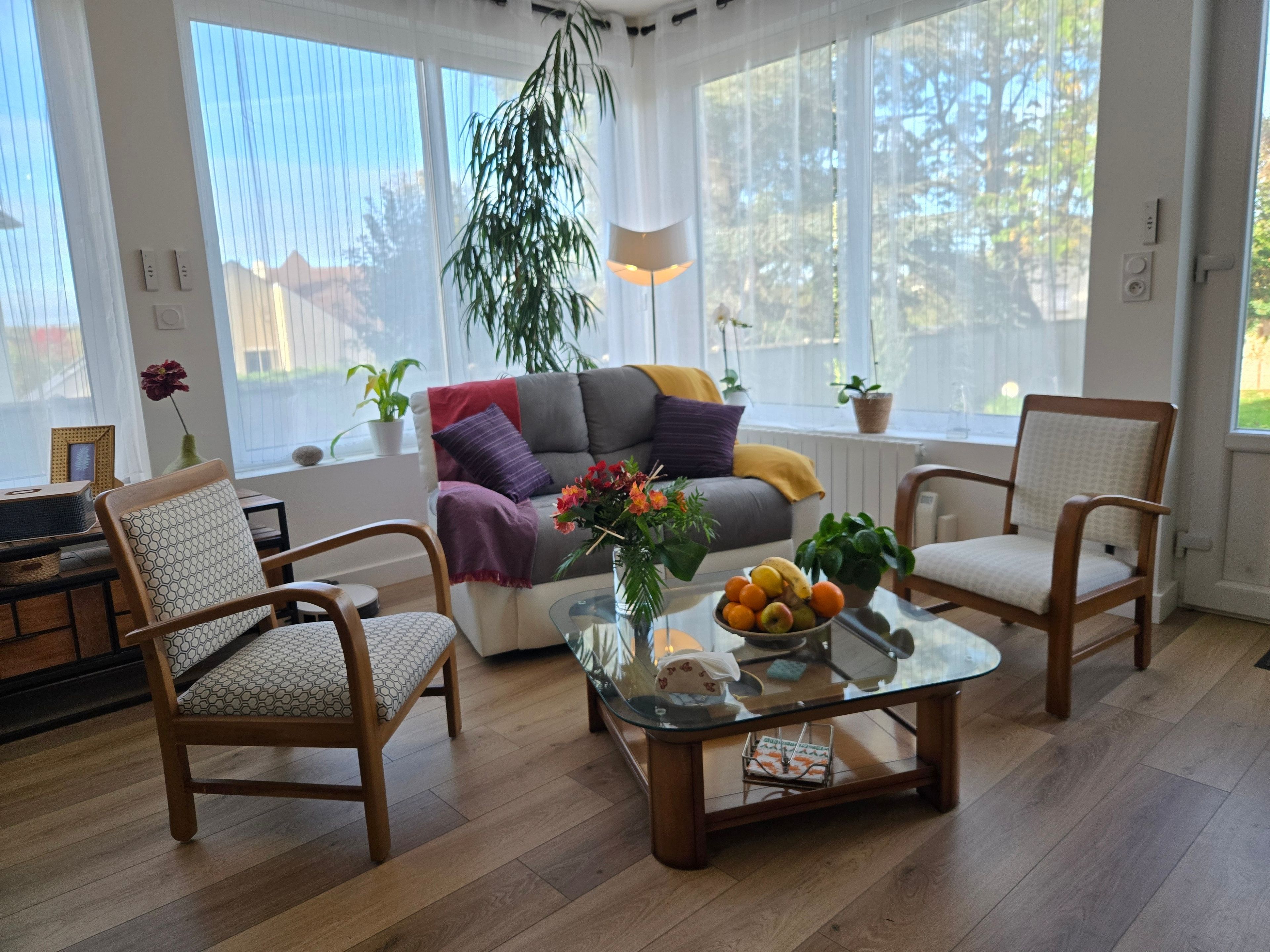 Un salón luminoso con un sofá, dos sillones, una mesa de centro con fruta y una planta colgante.