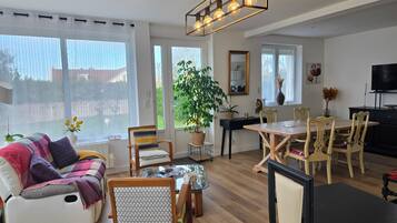 A modern living room with a sofa, dining table, chairs, a hanging light fixture, and a large window with curtains.