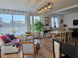 A modern living room with a sofa, dining table, chairs, a hanging light fixture, and a large window with curtains.