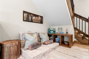 Um quarto acolhedor com um banco de madeira, um banquinho estofado com almofadas decorativas, um banquinho de madeira e uma estante com livros e um candeeiro.
