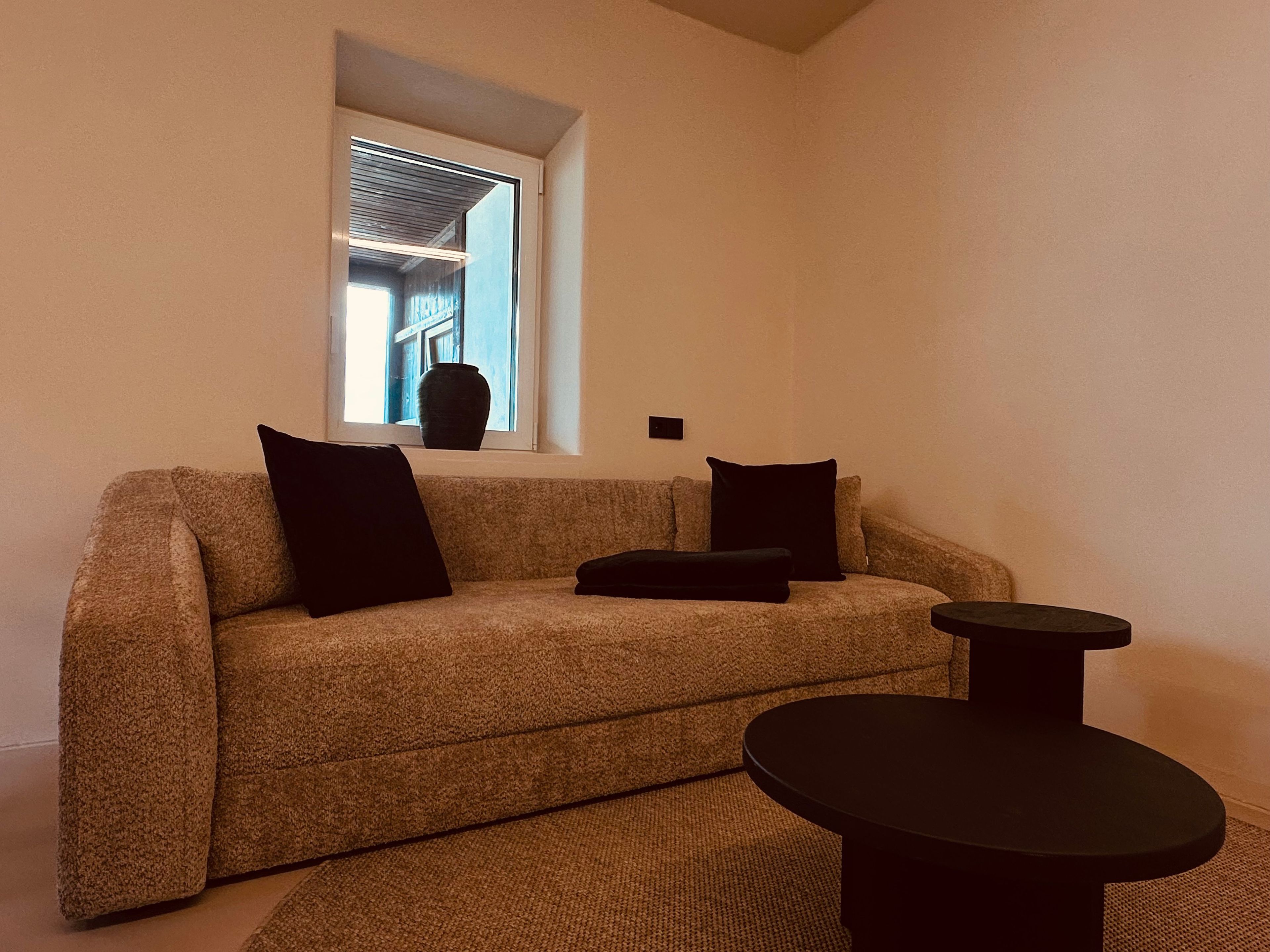 Un salon avec un canapé, deux coussins noirs, une petite table et une fenêtre donnant sur un immeuble.
