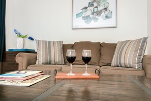 A living room with a brown sofa, striped pillows, a coffee table with two wine glasses, and a framed abstract painting on the wall. - Cozy 1BR retreat with king bed, WiFi, fire pit, and walkable Fredericksburg charm. (Fredericksburg)