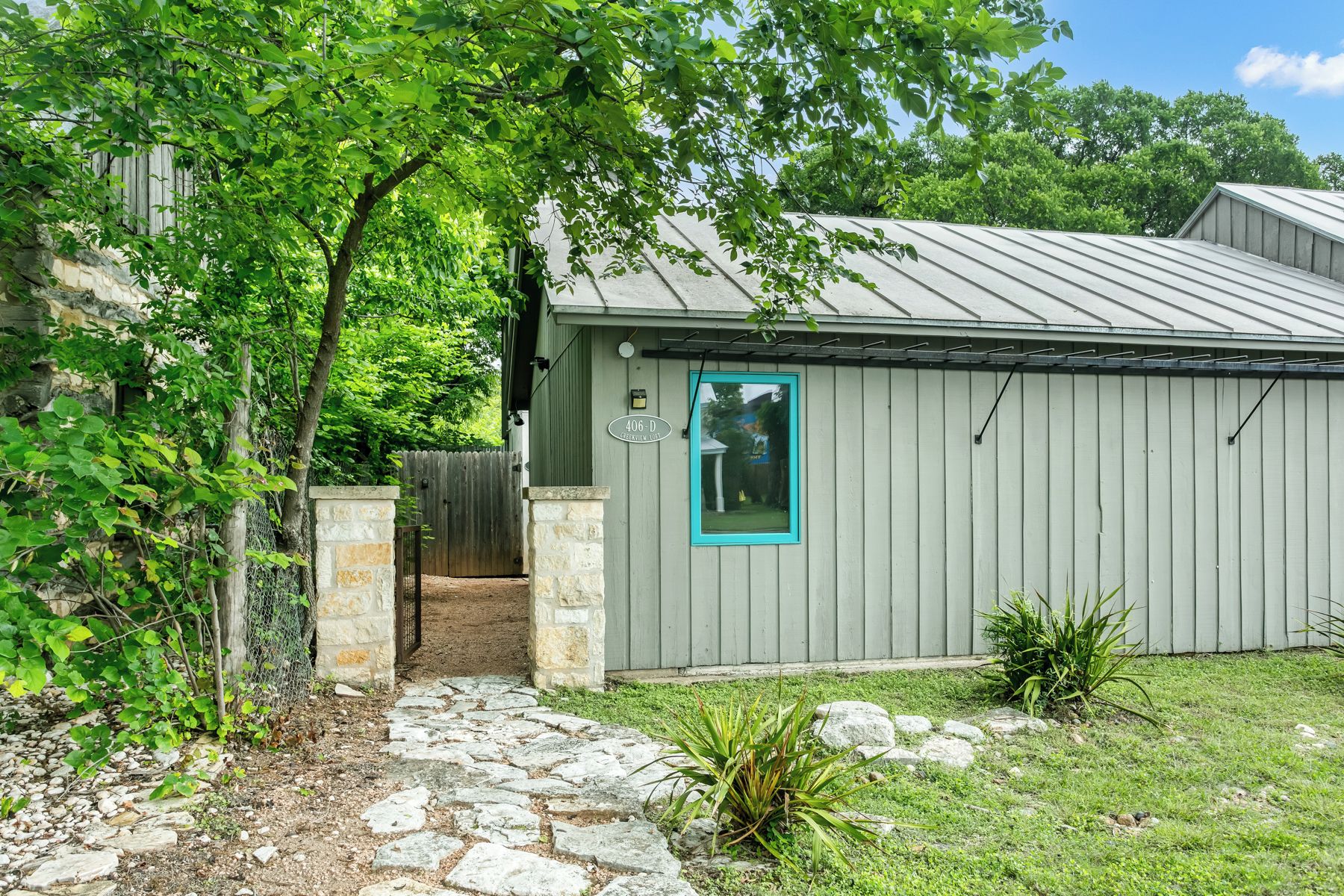 A building with a stone pathway leading to a wooden gate, surrounded by greenery and a metal roof.