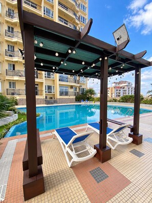 A pool area with lounge chairs, a pergola with string lights, and a building with balconies in the background. - Spacious and modern 2-bedroom apartment in Central Osu, Chateau Apartments (Accra)