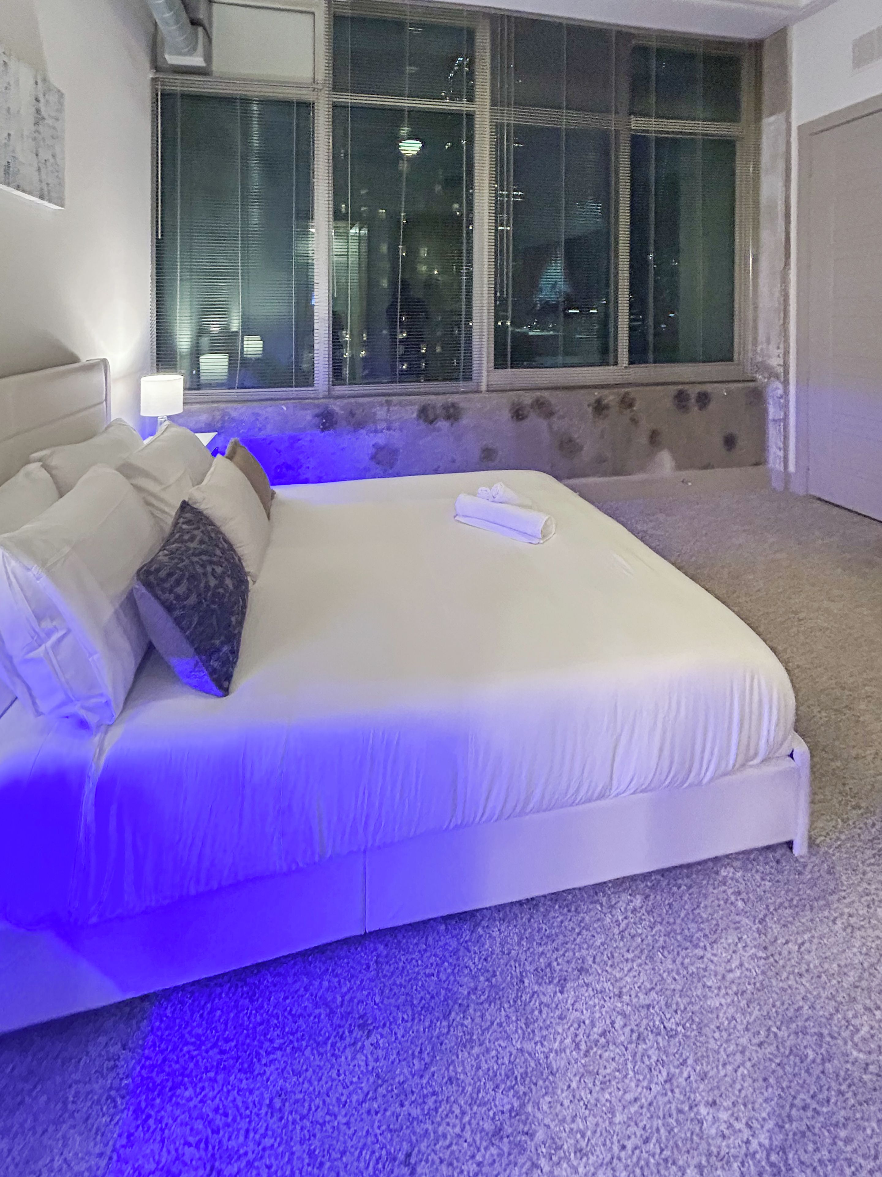 A neatly made bed with a headboard, a window with blinds, and a carpeted floor.