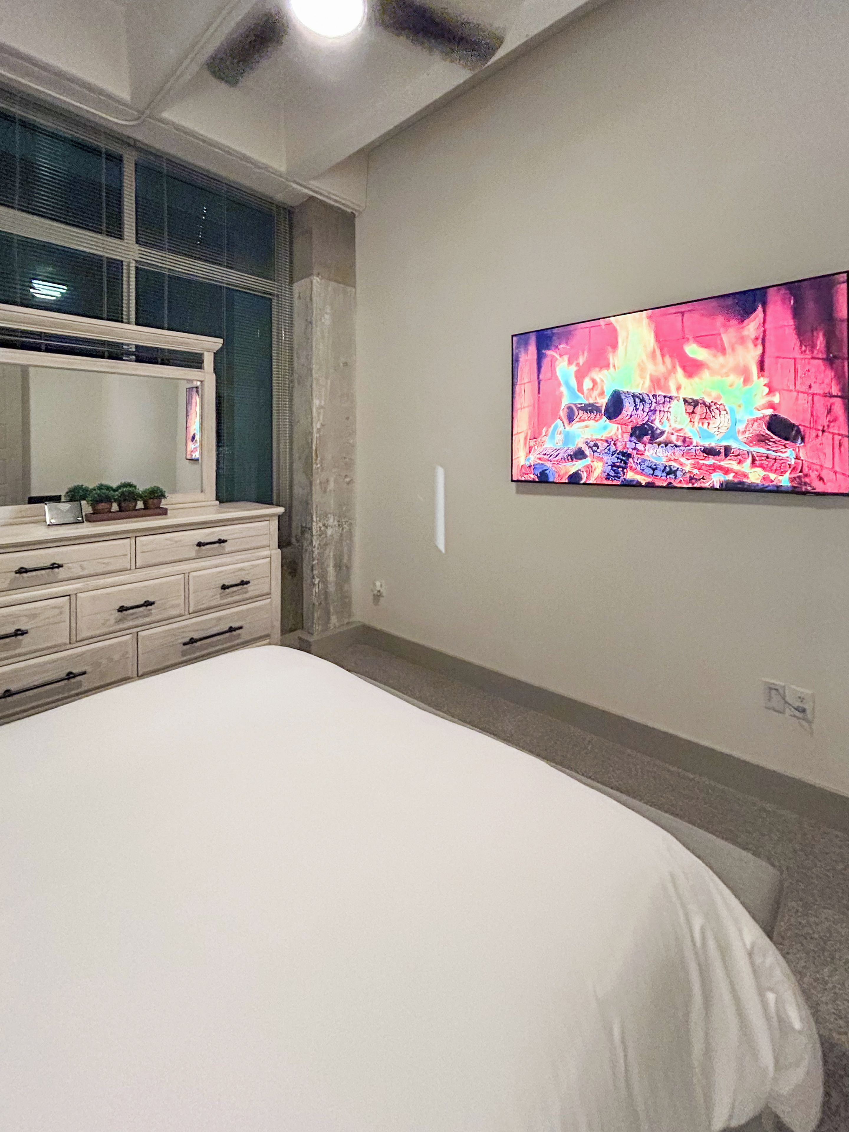 A bedroom with a bed, a dresser, a large mirror, and a wall-mounted TV displaying a fire scene.