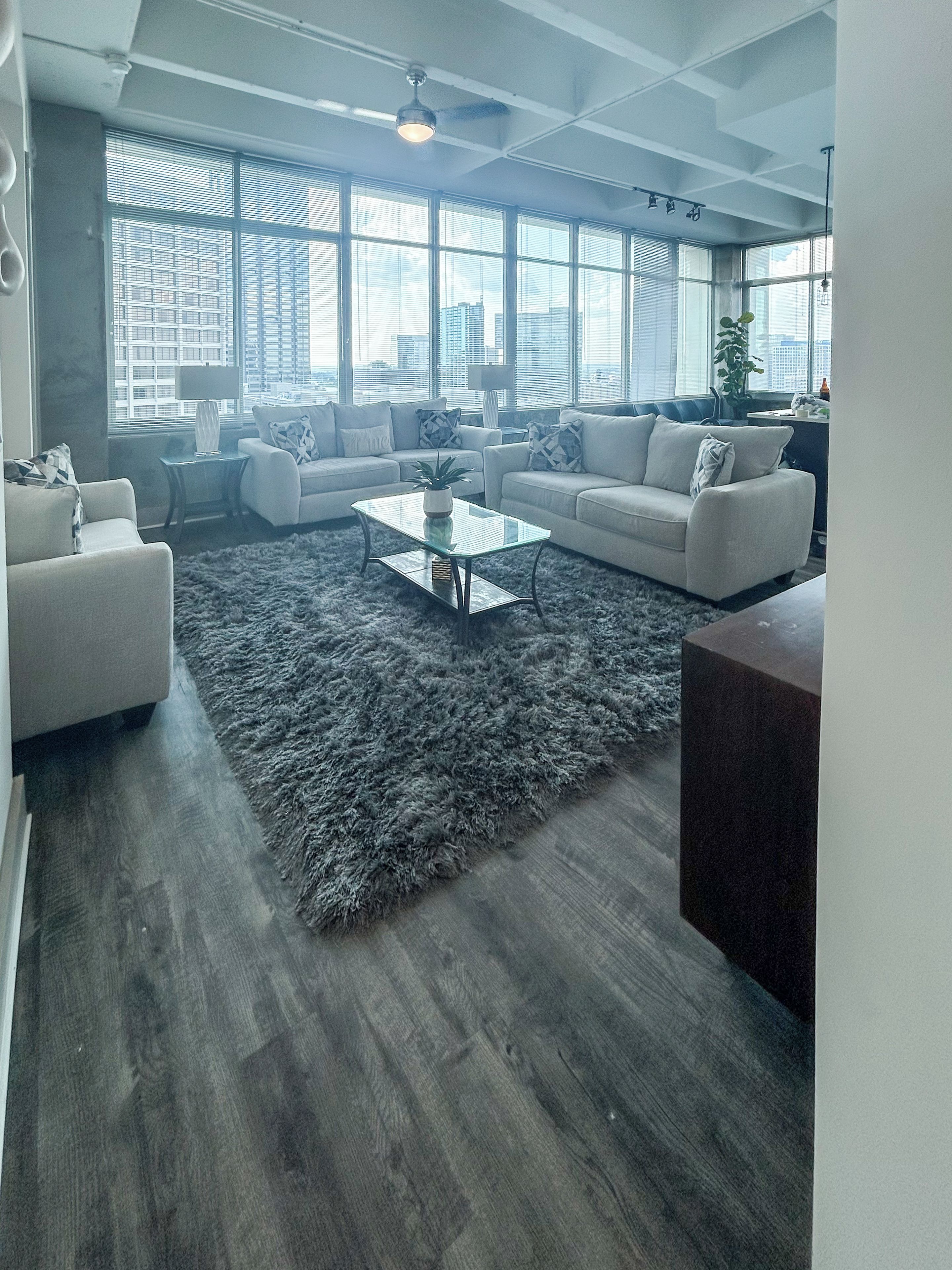 A modern living room with a large window, a glass coffee table, and a grey shag rug.