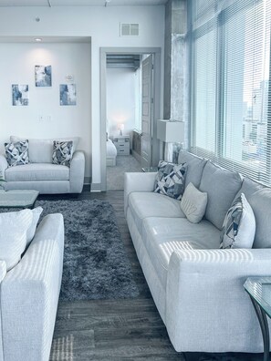 A modern living room with two sofas, a coffee table, and a rug. There is a door leading to another room and a window with blinds. - Altitude Apartments (Atlanta)