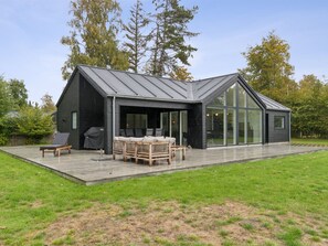 Et moderne hus med en stor glasfacade, en terrasse med udendørs møbler og en velplejet græsplæne.