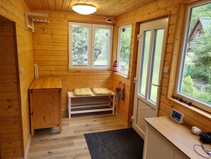 Interior de una cabaña de madera con un banco, una mesa pequeña y una ventana con vista a la vegetación.