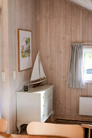 A room with wooden walls, a white dresser with drawers, a small sailboat model, a framed picture, and a window with curtains. - Summer house with two terraces near Rødhus Strand (Pandrup)