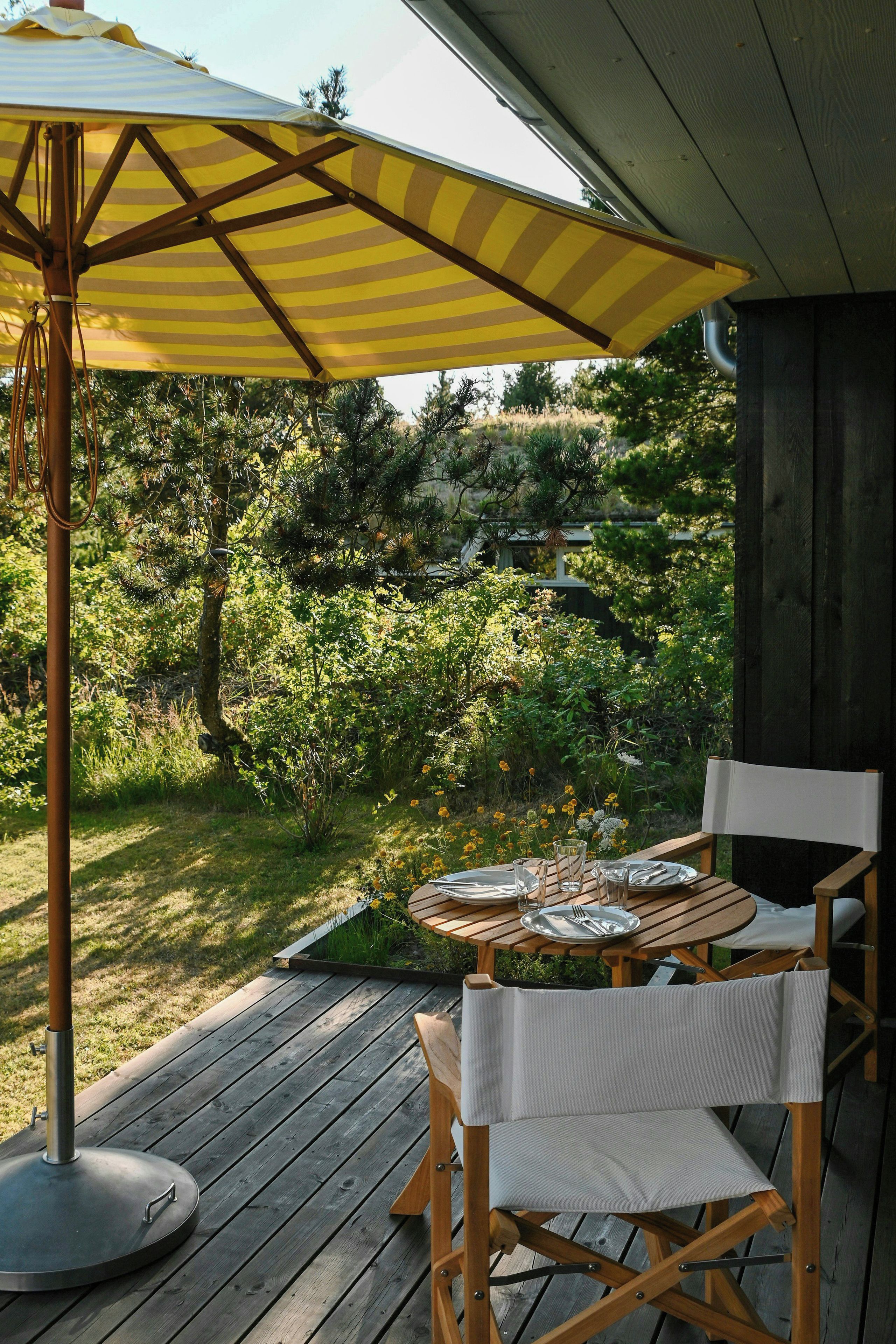 Un espace de restauration en plein air avec une terrasse en bois, une table dressée pour deux personnes et un parasol jaune à rayures offrant de l’ombre.